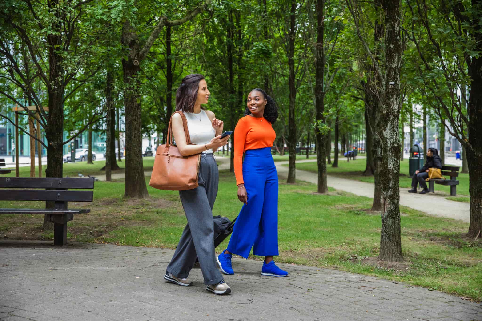 Two women walking in the park, smiling and talking with suitcase and phone. 