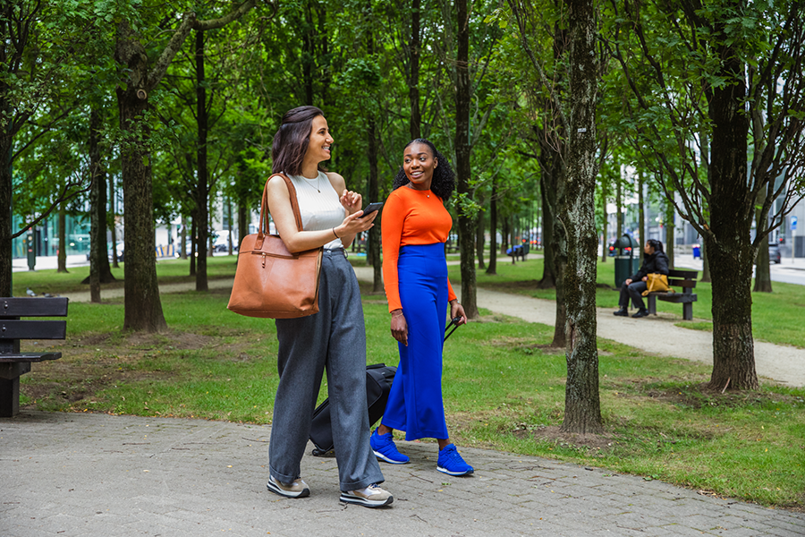 Två kvinnor som går i parken, ler och pratar med resväska och telefon.